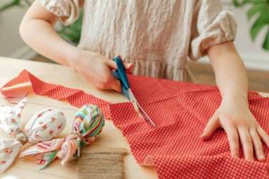 A girl at home makes Easter egg decorations in the shape of a rabbit from fabric. The child cuts the fabric with scissors. Ideas for home decoration.