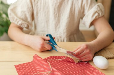 A girl at home makes Easter egg decorations in the shape of a rabbit from fabric. The child cuts the fabric with scissors. Ideas for home decoration.