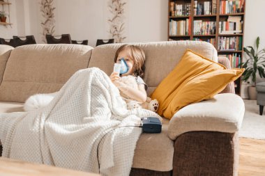 Little boy makes inhalation with a nebulizer at home lying on the couch. Cough prevention and treatment at home.
