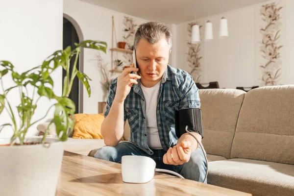 A man measures blood pressure at home with a blood pressure cuff and a tonometer. A man calls a doctor on a mobile phone. Health check at home. Consultations with a doctor online.
