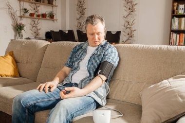 Evde adamın biri tansiyonunu kelepçeli bir tansiyon monitörüyle ölçüyor. Evde sağlık kontrolü. Hipotansiyon veya hipertansiyon konsepti.