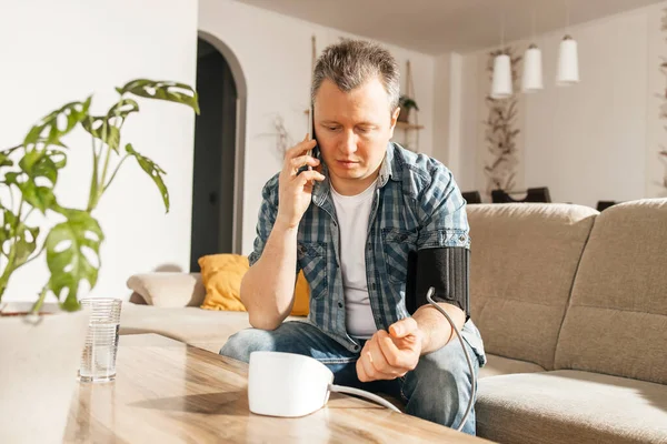A man measures blood pressure at home with a blood pressure cuff and a tonometer. A man calls a doctor on a mobile phone. Health check at home. Consultations with a doctor online.