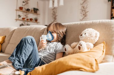 Little boy makes inhalation with a nebulizer at home lying on the couch. Cough prevention and treatment at home.