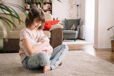 Genç bir kız oturma odasında yerde oturuyor ve sevgili kediciğiyle oynuyor. Çocuk evcil hayvana sarılıyor ve okşuyor. Çocuklar ve kediler