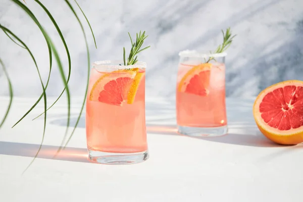 グレープフルーツと氷のフレッシュで冷たい夏のカクテルを備えた2つのグラスは ローズマリーと新鮮なフルーツスライスの小枝を添えています 赤オレンジドリンク 晴れた日の影 — ストック写真