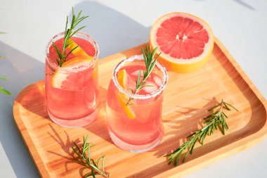 Güneşli bir günde, gri bir arka planda, ahşap servis tepsisinde soğuk yaz kokteylleri, kırmızı portakal, biberiye ve buz. Tatil kavramı