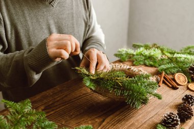 Bir adam evi için taze ladin dalları ve kurutulmuş portakal dilimlerinden Noel süsü yapar. DIY ev dekorasyonu.