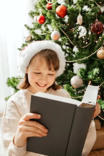 Noel Baba şapkalı mutlu bir kız süslü bir Noel ağacının yanında oturur ve kitap okur. Yılbaşını ve Noel 'i bekliyorum..