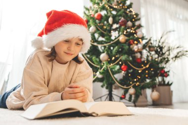 Noel Baba şapkalı mutlu bir kız süslü bir Noel ağacının yanında oturur ve kitap okur. Yılbaşını ve Noel 'i bekliyorum..