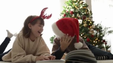Noel Baba şapkalı çocuklar yeni yıl ağacının önünde oturma odasında yerde yatarken kitap okuyup gülüyorlar.. 
