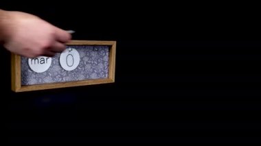 A wooden calendar block showing the date March 3rd with a mans hand putting on and taking off the metal discs with the date and month on them, filmed in 8k quality