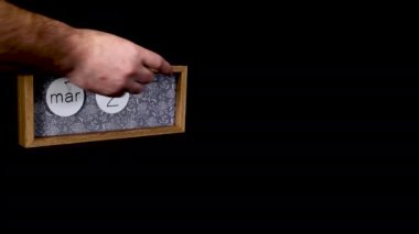 A wooden calendar block showing the date March 24th with a mans hand putting on and taking off the metal discs with the date and month on them, filmed in 8k quality