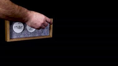 A wooden calendar block showing the date March 25th with a mans hand putting on and taking off the metal discs with the date and month on them, filmed in 8k quality
