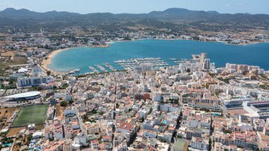 İspanya 'nın Balear Adaları' ndaki İbiza adasındaki Sant Antoni de Portmany kasabasındaki bir kumsalın insansız hava aracı fotoğrafı tekne limanını ve Playa de San Antonio olarak bilinen plajı gösteriyor.