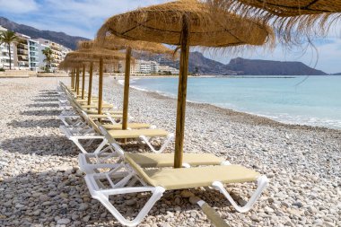 Altea 'daki güzel plajın, İspanya' daki Alicante 'nin fotoğrafı Playa La Roda olarak bilinen çakıl taşı plajında güneşlik ve saman plaj şemsiyelerini gösteriyor.