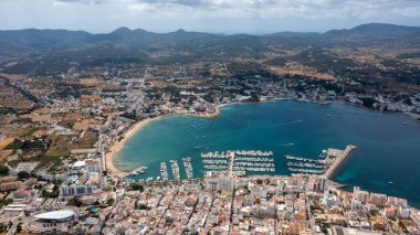 İspanya 'nın Balear Adaları' ndaki İbiza adasındaki Sant Antoni de Portmany kasabasındaki bir kumsalın insansız hava aracı fotoğrafı tekne limanını ve Playa de San Antonio olarak bilinen plajı gösteriyor.