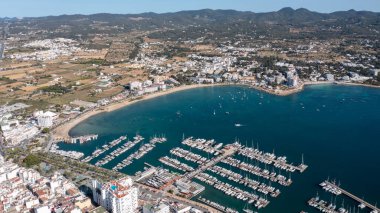 İspanya 'nın Balear Adaları' ndaki İbiza adasındaki Sant Antoni de Portmany kasabasındaki bir kumsalın insansız hava aracı fotoğrafı tekne limanını ve Playa de San Antonio olarak bilinen plajı gösteriyor.
