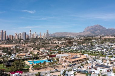 İspanya 'daki Benidorm kasabasının hava fotoğrafı, güneşli bir günde arka planda dağlar ve daireler olan kamp alanında birçok karavan ve karavanın bulunduğu bir kamp alanının insansız hava aracını gösteriyor.