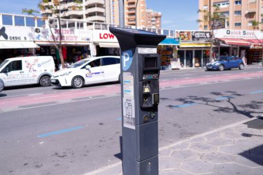 Benidorm İspanya, 4 Nisan 2024: Güneşli bir yaz gününde İspanya 'nın Benidorm kentindeki bir parkmetrenin fotoğrafı park cezası makinesinin ve trafiğin arkadan geçişini gösteriyor
