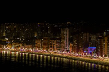 Benidorm İspanya, 5 Temmuz 2024: Benidorm Playa Levante plajının gece fotoğrafı, güneşin dağların ve otellerin arkasında batışını ve plajdaki yüksek katlı daireleri gösteriyor.