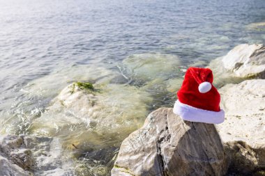 Photo on the beach of Sitio de Calahonda in Spain showing a Christmas festive Santa hat on a rock by the ocean indicating a Christmas by the sea concept clipart