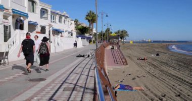 Benalmadena, Malaga, İspanya, 10 Ocak 2025: İspanya 'nın Benalmadena kasabasındaki güzel sahil önü ve sahil yürüyüşünün yavaş çekim görüntüleri, güneşli bir günde yürüyen genç bir çifti gösteriyor