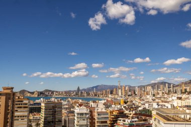İspanya 'nın eski Benidorm kasabasına bakan fotoğraf, arka planda beyaz bulutlar ve yüksek katlı apartmanlar ve binalarla çevrili Batı Sahili Gezinti Alanı' nı gösteriyor.