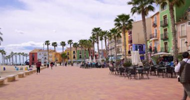 Villajoyosa, Valencia İspanya, 12 Mart 2025: İspanya 'nın Villajoyosa kentindeki palmiye ağaçları ve restoranları ile caddeyi gösteren tipik bir İspanyol caddesinin yavaş çekim görüntüleri