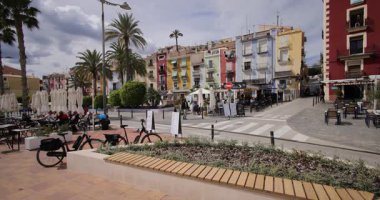 Villajoyosa, Valensiyalı İspanya, 12 Mart 2025: Villajoyosa kasabasındaki tipik bir İspanyol caddesinin görüntüsü palmiye ağaçları ve renkli restoranların bulunduğu caddeyi gösteriyor