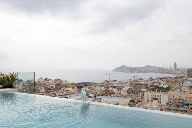 Sıcak ama bulutlu bir günde, arka planda İspanya 'nın Benidorm kasabasıyla birlikte, yüzme havuzunun kenarında oturan beyaz bir martı kuşunun fotoğrafı.
