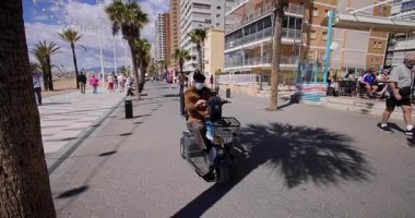 Benidorm İspanya, 27. Maç 2025: İspanya 'nın Benidorm kasabasının yavaş çekim görüntüleri büyük sahil cephesinde yürüyen insanları ve hareket halindeki scooter' da maske takan bir adamı gösteriyor.