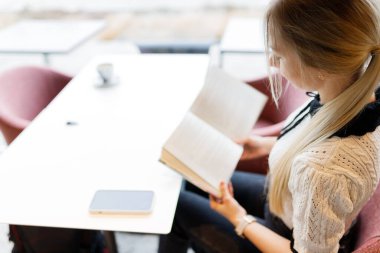 Genç bir kız kafede bir fincan kahve eşliğinde kitap okuyor..