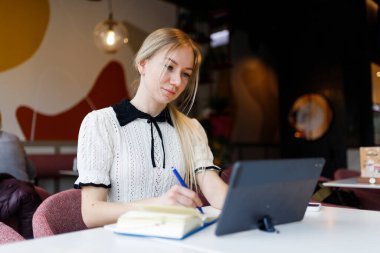 Kız uzaktan tablet üzerinde çalışıyor ve bir not defterine bilgi yazıyor.