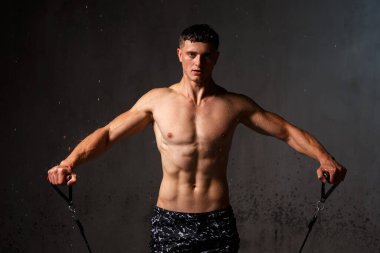 Vücut geliştirici stüdyoda. Dambılları ve gri arka planda çıplak gövdesi olan sandalyeleri var..