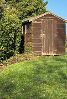 Asma kilitli kapısı olan eski ahşap baraka, üzerinde mavi gökyüzü ve çimenlerde yan yana çalılar.