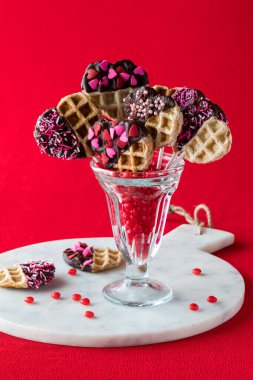 Tarçınlı bir kalp, içinde süslü sevgililer günü waffle çubukları bulunan tatlı bardağını kırmızı arka planda tutuyor..