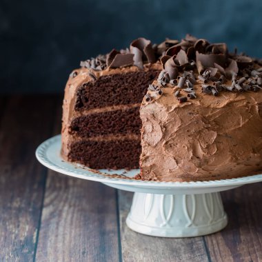 A triple layered moist chocolate cake with frosting and chocolate curls on a pedestal stand against a dark background.