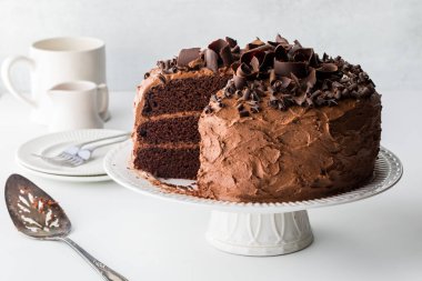 A close up of a triple layered chocolate cake with one slice removed, against a light background.