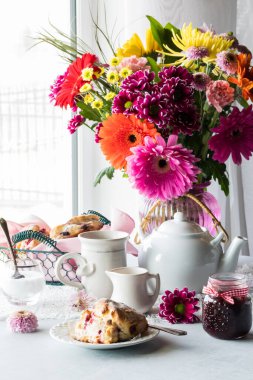 Arkasında güzel bir çiçek buketi olan çay ve çörekler, parlak güneşli bir pencereye karşı..