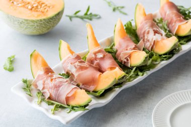 Prosciutto ambalajlı kavun dilimlerinin üstünde bir tabak roka. 