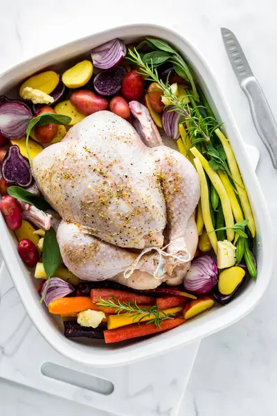 A raw whole chicken surrounded by raw colourful vegetables, ready for baking.