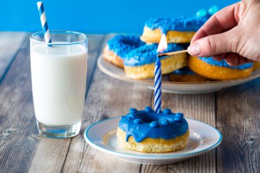 Bir el, arkasında bir tabak donut olan ve sütle servis edilen mavi donutun içine yerleştirilmiş bir mum yakıyor..