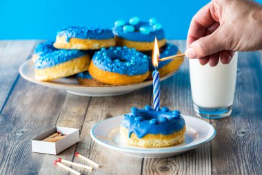 Bir el, arkasında donutlarla dolu mavi çikolatalı bir çöreğin üzerinde mum yakıyor..
