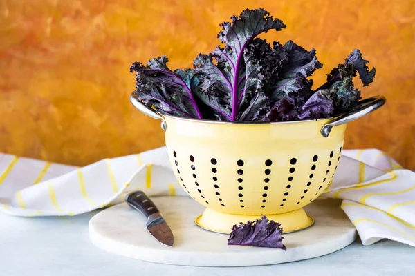 A close up of fresh purple kale in a light yellow colander against an orange background.