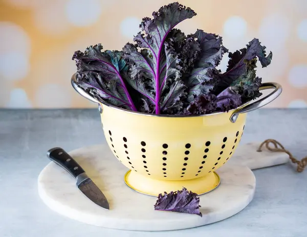 A close up of fresh purple kale in a yellow colander, on a marble slab.