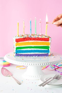 Gökkuşağı pastasının üstünde mum yakan bir elin yakın çekimi..