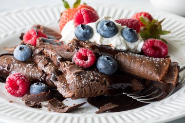 A close up of chocolate crepes topped with whipped cream, berries and chocolate sauce.  clipart