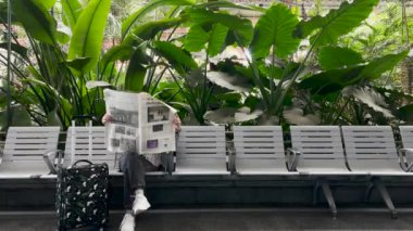 Ukraynalı bir mülteci kadın bekleme bankında oturuyor ve New York Times gazetesini elinde bavulla tropikal palmiye ağaçları olan bir tren istasyonunda okuyor. İspanya Madrid