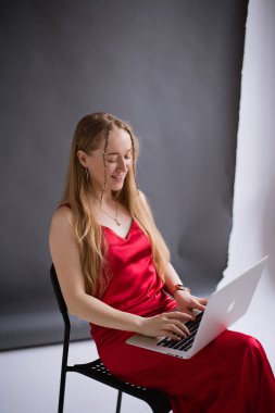 Kırmızı elbiseli mutlu bir iş kadını blogger gülüşü işe yarıyor, dizüstü bilgisayarda bir elma yazıyor ve bir sandalyede oturuyor. Bilgisayar MacBook 'lu sarışın asistanın portresi. Dikey