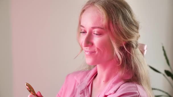 自然な金髪の女性は ピンクのネオン照明でブラシでバービーメイクをしている クローズアップ カメラを見ています 花嫁は粉を適用し 彼女の顔にブラシをかけ 前に独身パーティーの準備をする — ストック動画